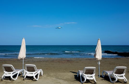 Sun beds and beach umbrellas on the beach of the Mediterranean sea against the background of a plane coming in for landing.