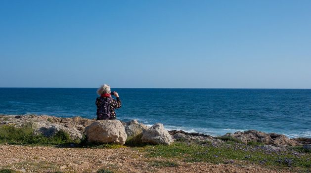 An elderly woman with gray hair takes photos on her phone of waves on the beach on a Sunny, windy day.