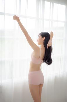 Beautiful young asian woman waking up in the morning stand stretching arms in the bedroom, sexy girl in underwear relax cozy and wake with fresh happy at home, lifestyle concept, back view.