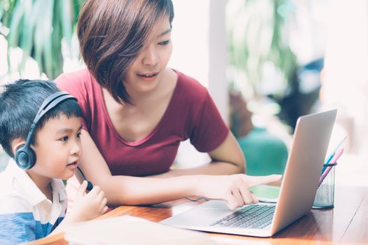 Young asian mother and son using laptop computer for study and learning together at home, boy wearing headphone for e-learning with distancing, teacher or mom support child, education concept.