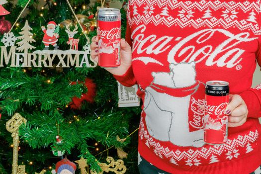 Woman wearing sweater with logo displays seasonal beverage before decorated tree.