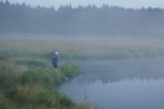 A fisherman in the fog catches fish on the pond.