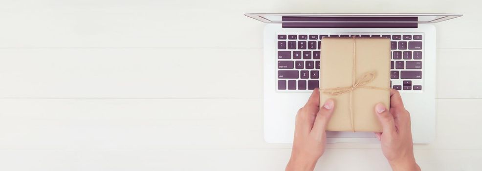 Hand of man holding gift box and having laptop computer on wooden table, present giving for Christmas day, season and celebrate, top view, flat lay, online shopping concept, banner website.