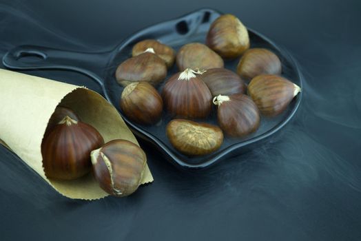 Roasting chestnuts in a black pan, surrounded by white smoke and a cone-shaped paper cone, a dish for a traditional healthy winter snack or appetizer