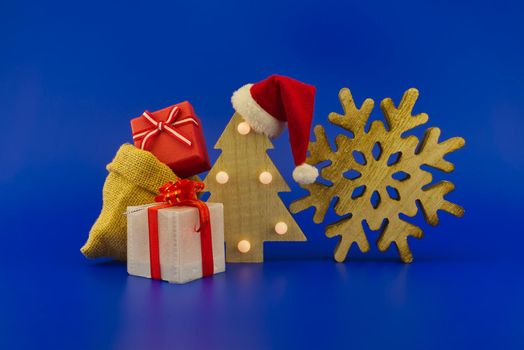 Stylized wooden Christmas tree with red Santa hat and jute sack with gift boxes on a festive blue background. New Year and Christmas gift season concept