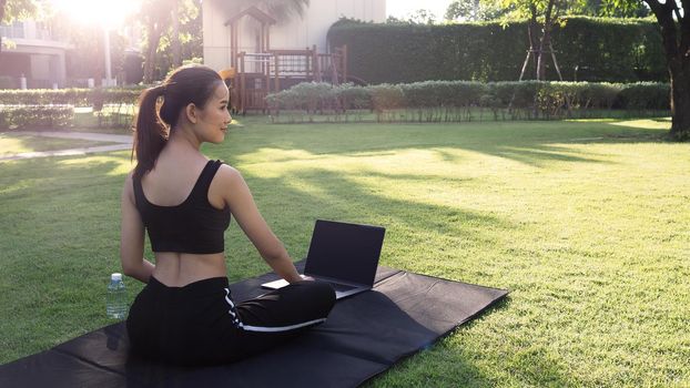 A sporty woman or online instructor in sportswear is sitting and practicing yoga on the mat with computer notebook and a bottle of water and is using a laptop at home in the backyard. Sport and recreation concept.