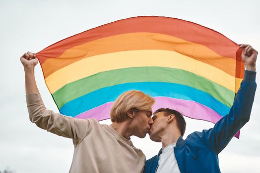 Romantic gay couple hugging, kissing and holding hands outdoors. Two handsome men holding LGBT pride flag.