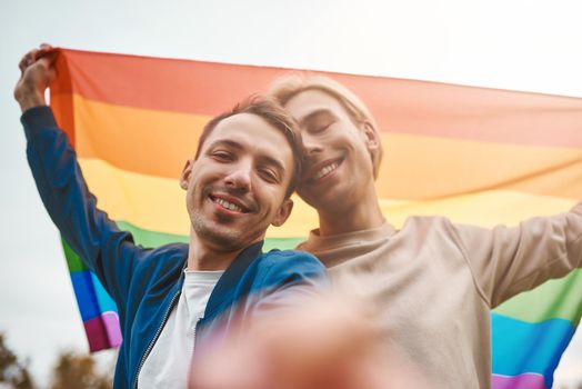 Romantic gay couple hugging, kissing and holding hands outdoors. Two handsome men holding LGBT pride flag while making selfie.