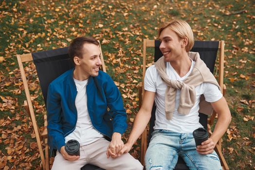 Loving gay couple sitting outdoors together and holding hands while drinking coffee. Two handsome men having romantic date in park. LGBT concept.