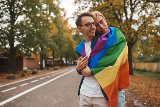 Romantic gay couple hugging, kissing and holding hands outdoors. Two handsome men holding LGBT pride flag in park.