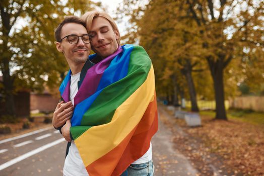 Romantic gay couple hugging, kissing and holding hands outdoors. Two handsome men holding LGBT pride flag in park.
