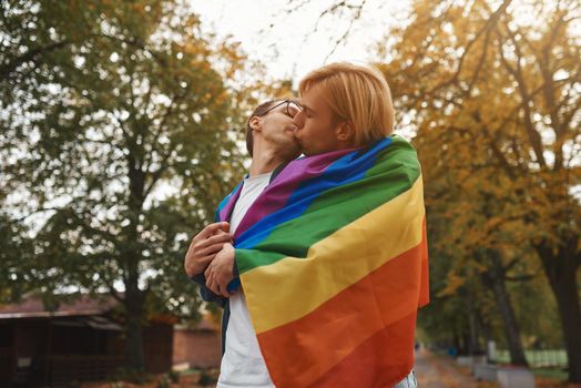 Romantic gay couple hugging, kissing and holding hands outdoors. Two handsome men holding LGBT pride flag in park.