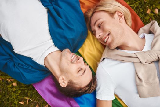 Loving gay couple having romantic date outdoors. Two handsome men lying together on LGBT pride flag.