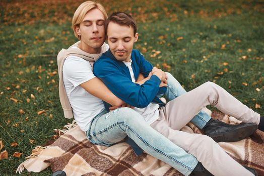 Loving gay couple having romantic date outdoors. Two handsome men sitting together on blanket in park and hugging. LGBT concept.