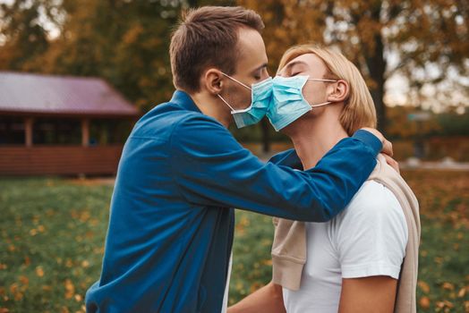 Romantic gay couple outdoors. Love during coronavirus. Two handsome men kissing wearing face masks. Covid-19 protection.