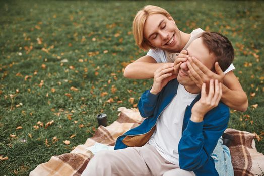 Loving gay couple having romantic date outdoors. Two handsome men sitting together on blanket in park. LGBT concept.