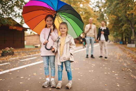 Two gay parents with their adopted daughters walking in park together. Two cute girls standing on foreground with rainbow umbrella. Happy LGBT family concept.