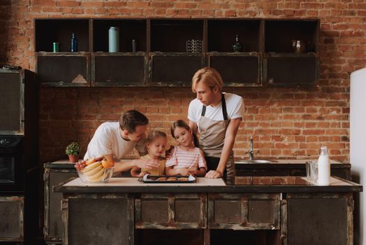 Gay couple with their adopted cute daughters cooking on kitchen. Lgbt family at home. Portrait of two handsome men and their two little daughters eating cookies.