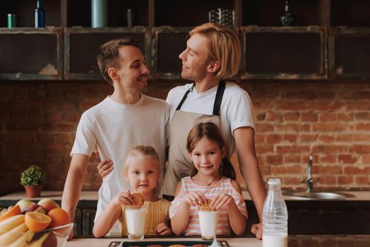 Gay couple with their adopted cute daughters cooking on kitchen. Lgbt family at home. Portrait of two handsome men and their two little daughters eating cookies and drinking milk.