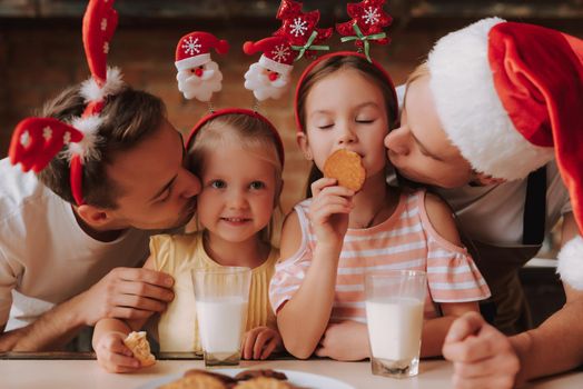 Gay couple with their adopted cute daughters cooking on kitchen. Lgbt family at home. Portrait of two handsome men and their two little daughters eating cookies and drinking milk wearing Santa Claus hat and other Christmas decoration. Christmas mood.