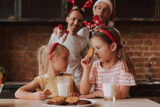 Gay couple with their adopted cute daughters cooking on kitchen. Lgbt family at home. Portrait of two handsome men and their two little daughters eating cookies and drinking milk wearing Santa Claus hat and other Christmas decoration. Christmas mood.