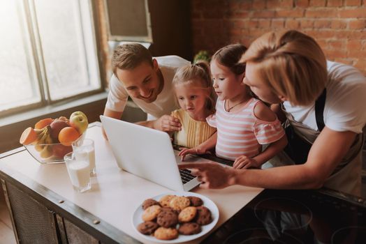 Gay couple with their adopted cute daughters reading recipe on laptop and cooking on kitchen. Lgbt family at home.
