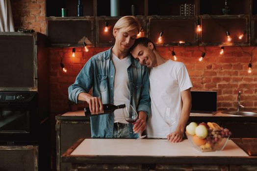 Loving gay couple at home. Two handsome men hugging and kissing on kitchen. Romantic atmosphere with bottle of red wine. LGBT concept.