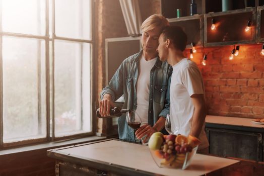 Loving gay couple at home. Two handsome men hugging and kissing on kitchen. Romantic atmosphere with bottle of red wine. LGBT concept.