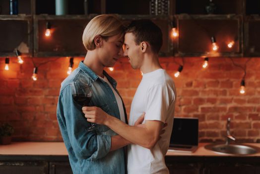 Loving gay couple at home. Two handsome men hugging and kissing on kitchen. Romantic atmosphere with bottle of red wine. LGBT concept.