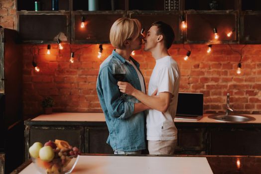 Loving gay couple at home. Two handsome men hugging and kissing on kitchen. Romantic atmosphere with bottle of red wine. LGBT concept.