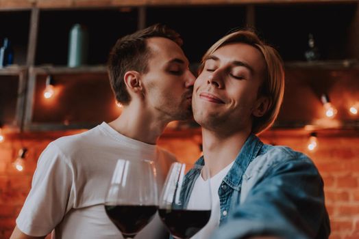 Loving gay couple at home. Two handsome men hugging, kissing and drinking wine on kitchen. LGBT concept.