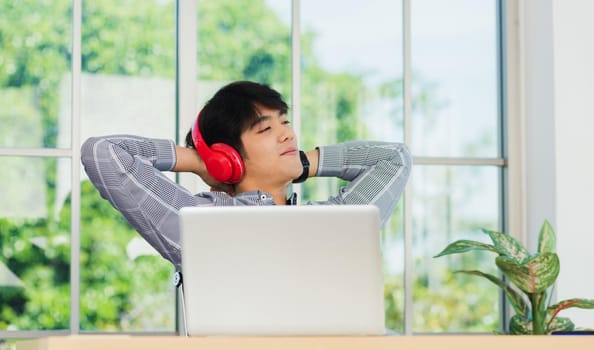 Asian young business man smile listening music in red headphone at home office. Happy businessman sitting on desk and relax wear wireless head phones enjoy listen music and work with laptop computer
