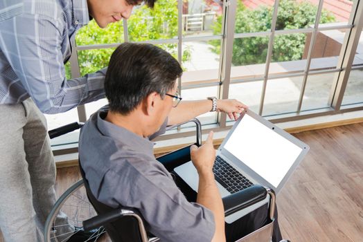 Asian senior disabled businessman in a wheelchair with laptop computer discuss together with team in office. Old father man sitting wheelchair and his son talking video calls conference on laptop