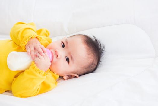 Asian beautiful little baby girl wearing a yellow dress eat milk sleeping feeding lying on the white bed, infant holding a bottle of milk, baby food concept