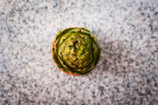 Green artichoke in a wooden bowl over a granite countertop. Top down view.