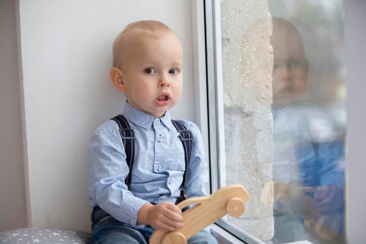 Little baby near the window. A kid with a wooden typewriter is sitting on a windowsill.