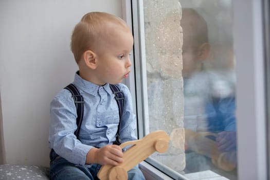 A small child looks out the window. The kid with a toy sits on a window sill.