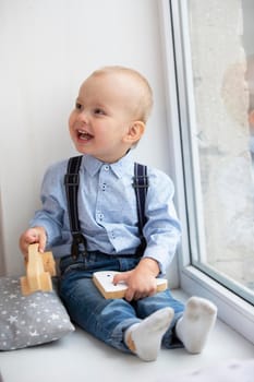 Little baby near the window. The cheerful kid with a toy sits on a window sill.
