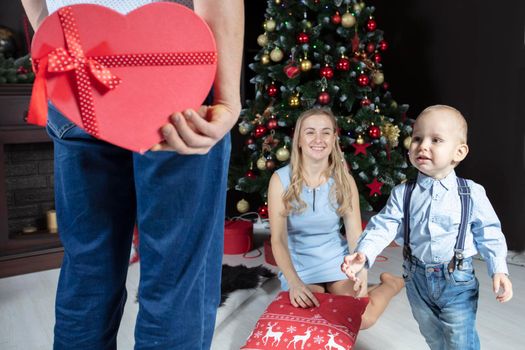 Christmas surprise. A man holds a red gift box on the background of his wife and child. Family on New Year's holidays. The little son runs to receive a Christmas gift.