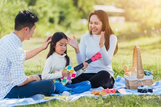 Happy Asian young family father, mother and children having fun and enjoying outdoor together sitting on the grass party with playing Ukulele during a picnic in the garden park on a sunny day