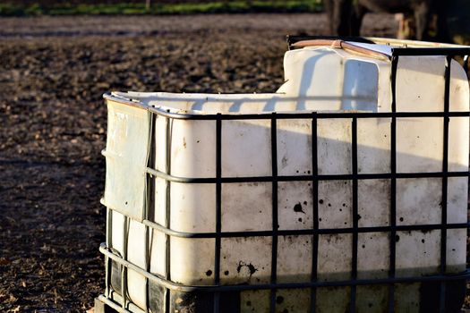 Big white water container with iron grid for stabilisation for animals on a paddock and the leg of a horse in the bachground