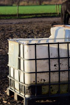 Big white water container with iron grid for stabilisation for animals on a paddock and the leg of a horse in the bachground