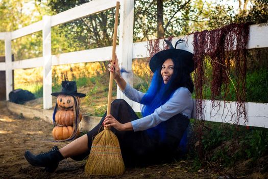 A girl dressed as a witch sits by a fence with a broom in her hands and shows her tongue