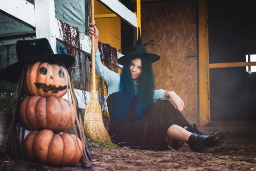 A girl dressed as a witch sits by a fence with a broom in her hands, in the foreground there is a scary figure of pumpkins