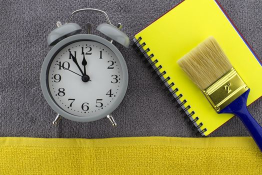 Color of the year and color trends themed still life with alarm clock placed to the side of yellow wire bound notepad and paintbrush on a textured grey background viewed as a flat lay from overhead