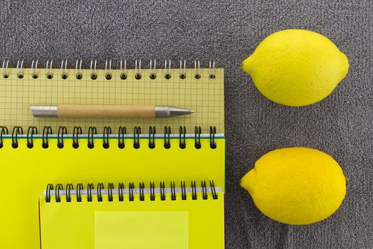 Colorful yellow themed still life with fresh healthy lemons placed to the side of three wire bound notepads and pen on a textured grey background viewed as a flat lay from overhead