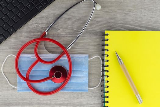 Coronavirus or Covid-19 medical concept with a colorful red stethoscope coiled over a surgical face mask alongside a notebook and pen on a wooden desk with computer keyboard viewed top down