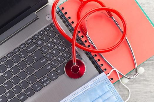 Coronavirus or Covid-19 medical concept with a colorful red stethoscope coiled over a surgical face mask alongside a notebook and pen on a wooden desk with computer keyboard viewed top down