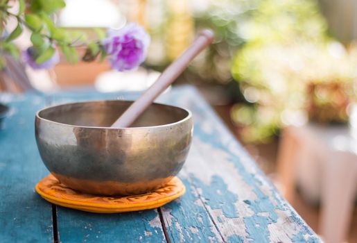 Metal singing bowl on a rustic green, wooden table outdoors. Flowers in the colourful, blurry background