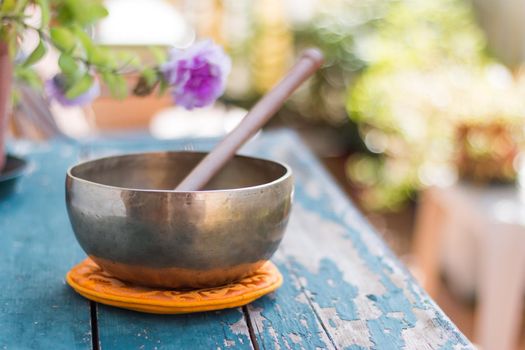 Metal singing bowl on a rustic green, wooden table outdoors. Flowers in the colourful, blurry background